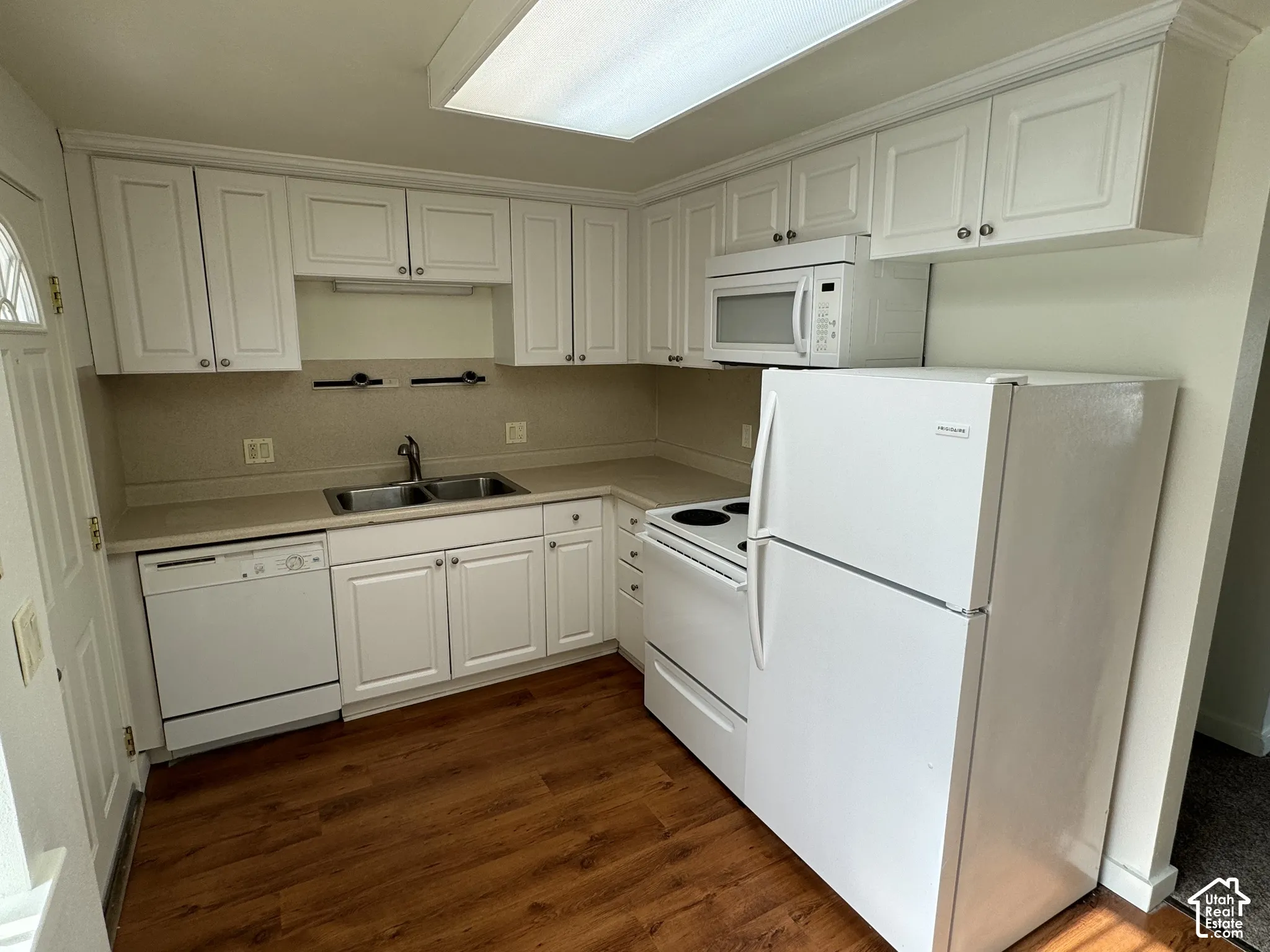 Kitchen featuring white cabinetry, light-toned countertops, a double basin sink, wood-finish flooring, and white appliances