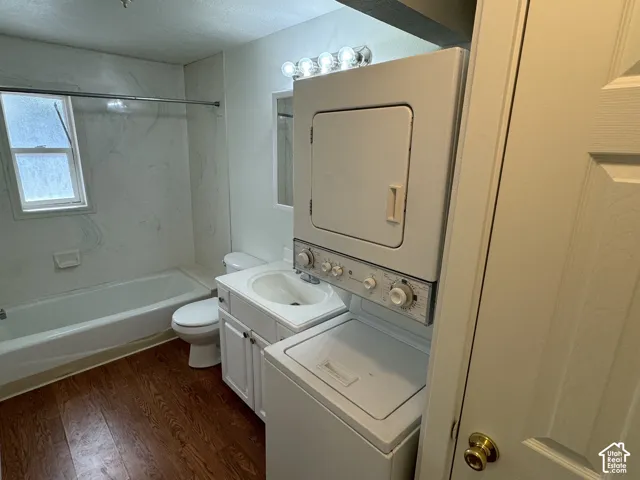 Wood-finish flooring, white vanity with integrated sink, toilet, tub with shower kit, and a stacked washer and dryer
