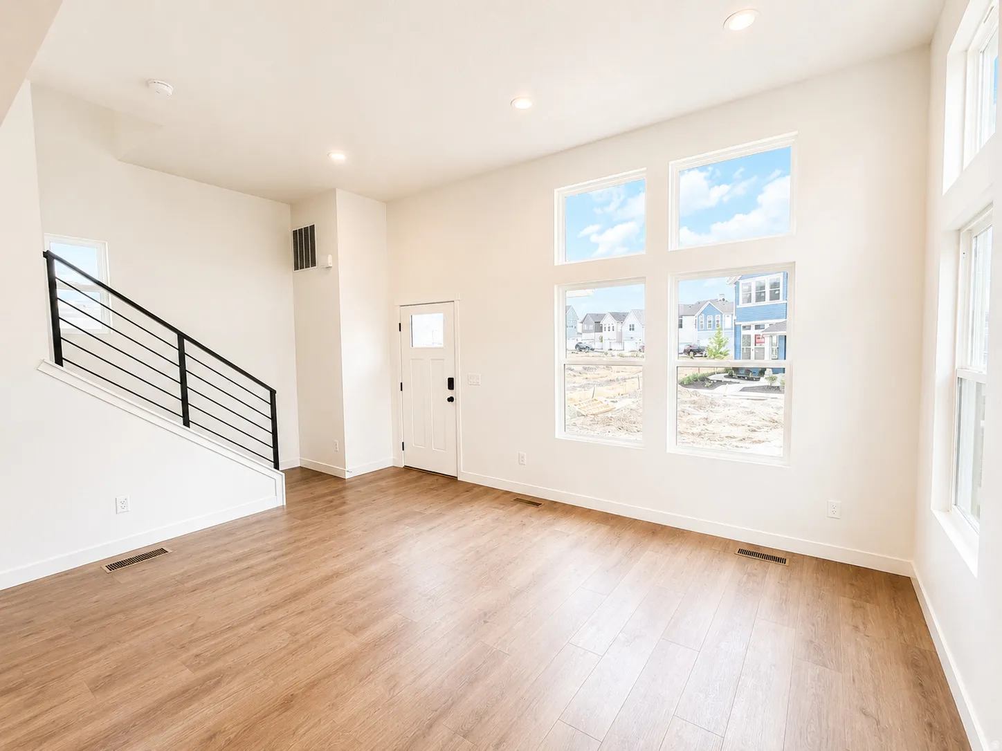 Expansive living area featuring high ceilings, wood-finish flooring, a modern black metal staircase railing, recessed lighting, and multiple large windows