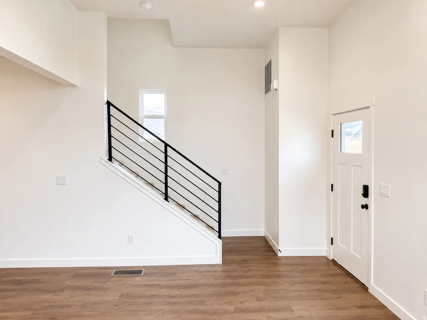 Wood-finish flooring throughout, bright white walls, modern staircase with black railing, recessed lighting, and a white entry door with a window pane