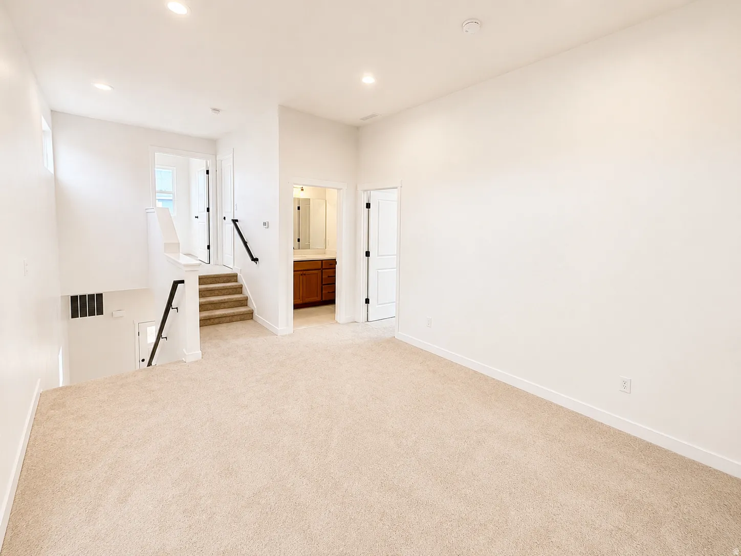 Expansive carpeted area with recessed lighting, crisp white walls, staircase with black metal railings, and a nearby bathroom featuring a wood-finish vanity with a mirror