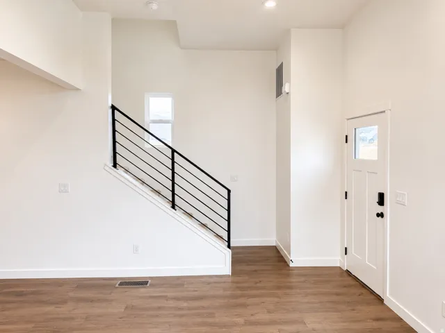 Wood-finish flooring throughout, bright white walls, modern staircase with black railing, recessed lighting, and a white entry door with a window pane