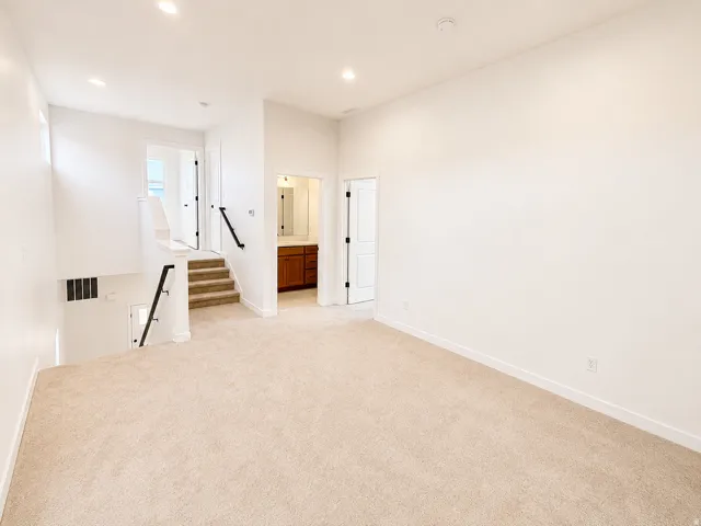 Expansive carpeted area with recessed lighting, crisp white walls, staircase with black metal railings, and a nearby bathroom featuring a wood-finish vanity with a mirror