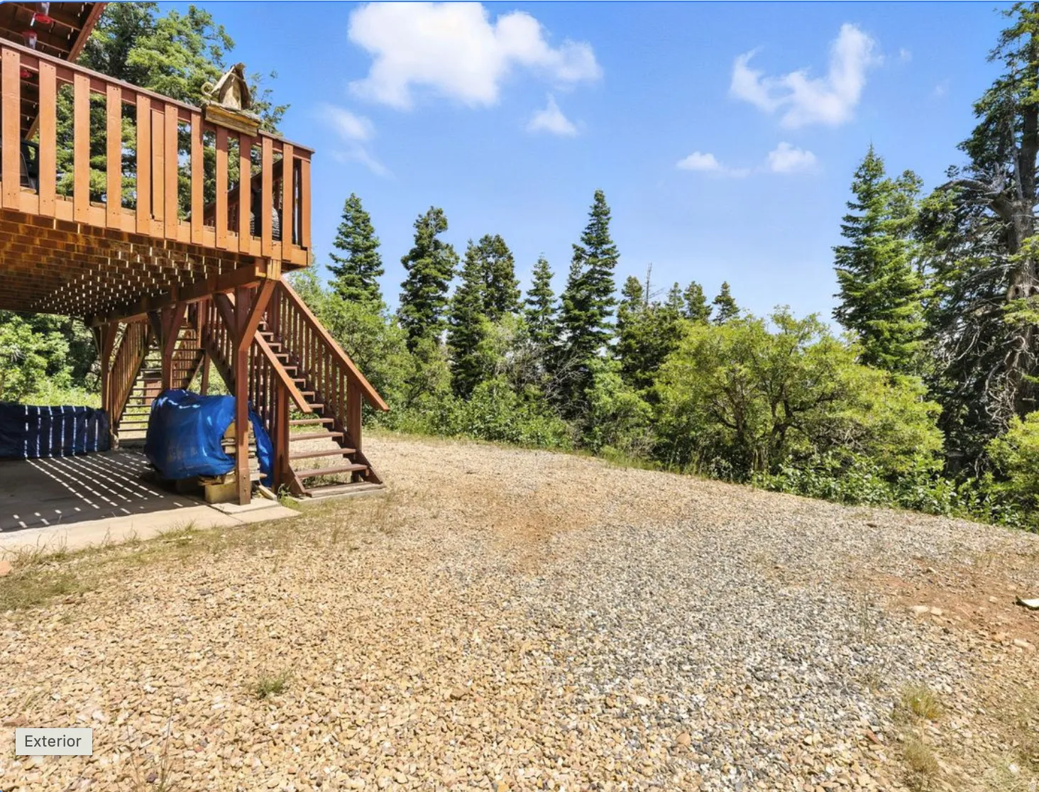 Expansive gravel yard featuring a wooden deck with railing, wooden support beams, and a wooden staircase