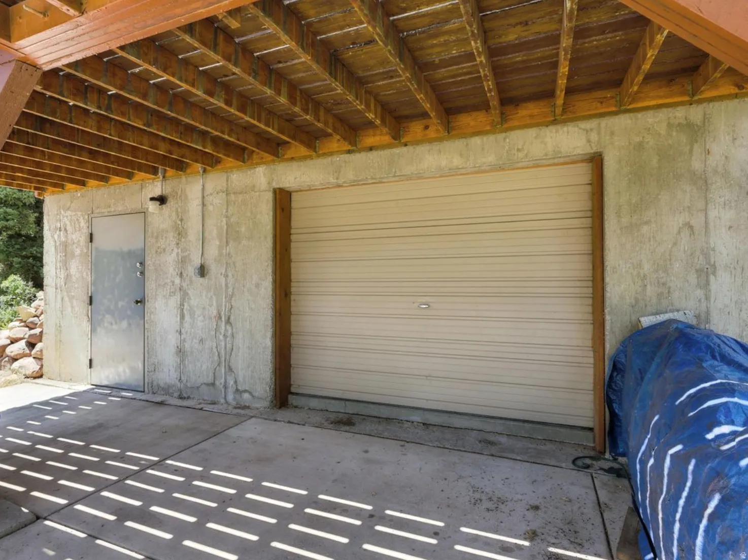 Under-deck parking area with a concrete slab, exposed wood joists, a corrugated garage door, and an exterior access door