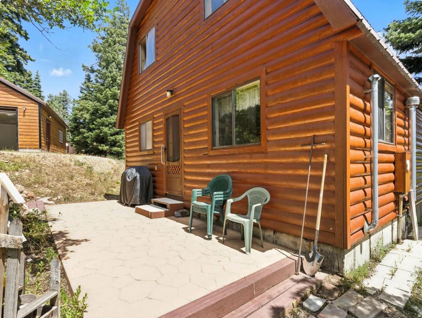 Log cabin exterior with a stone-paved patio, multiple windows, and a separate structure in the background