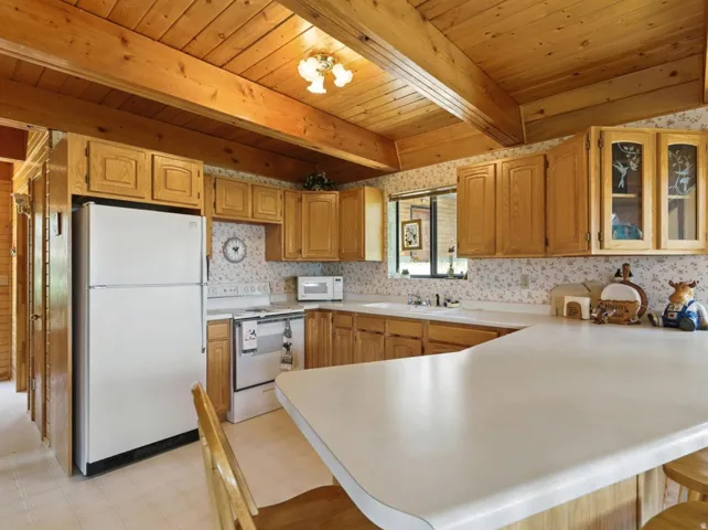 Wood-panel vaulted ceiling with exposed beams, wood-finish cabinetry, white countertops, built-in sink, and white appliances