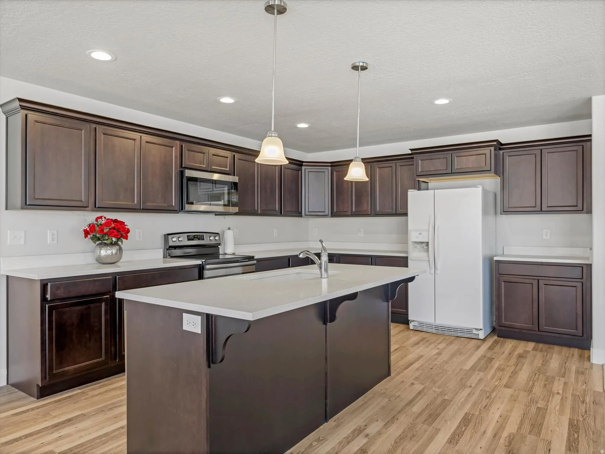Recessed lighting, wood-finish flooring, dark wood cabinetry, white countertops, and a kitchen island with a built-in sink