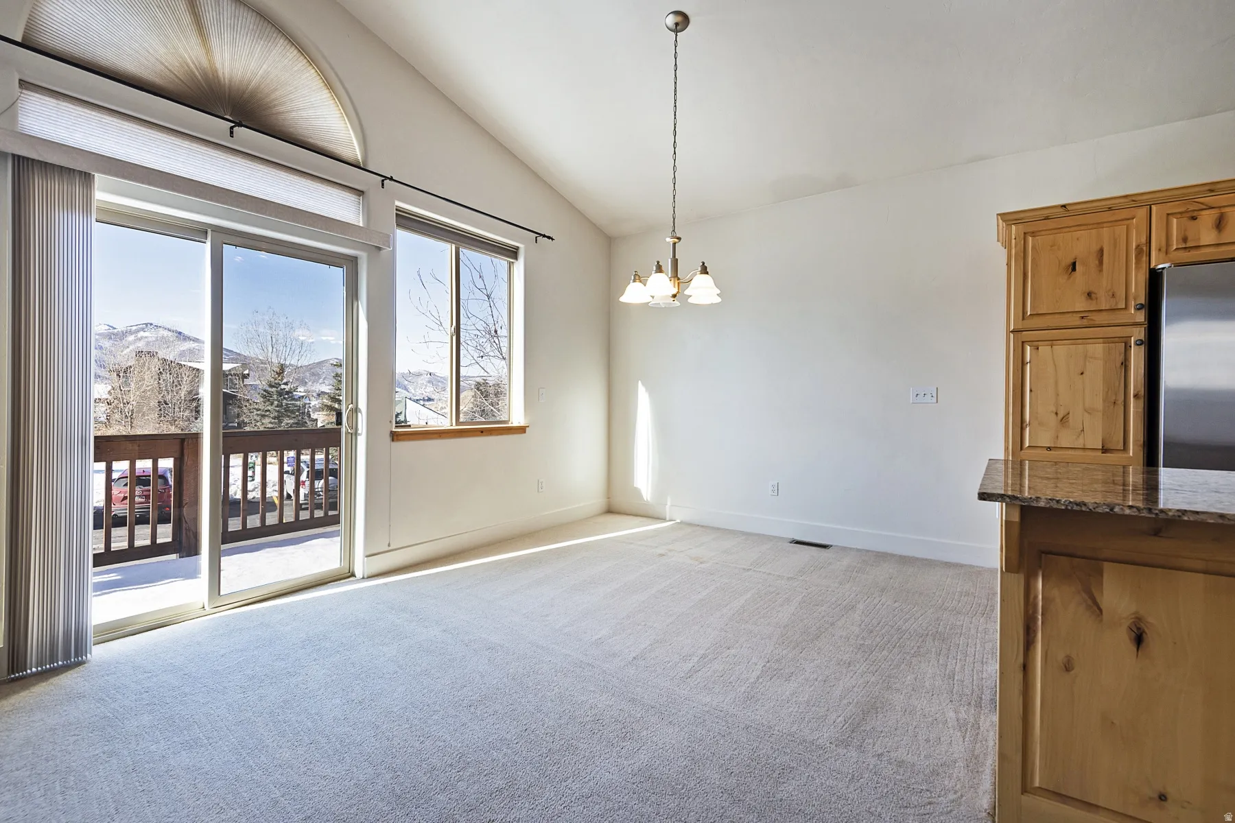 Vaulted ceilings with a semi-flush mount chandelier, sliding glass door to a balcony with wood railing, large windows, and built-in wood cabinetry with a stone countertop