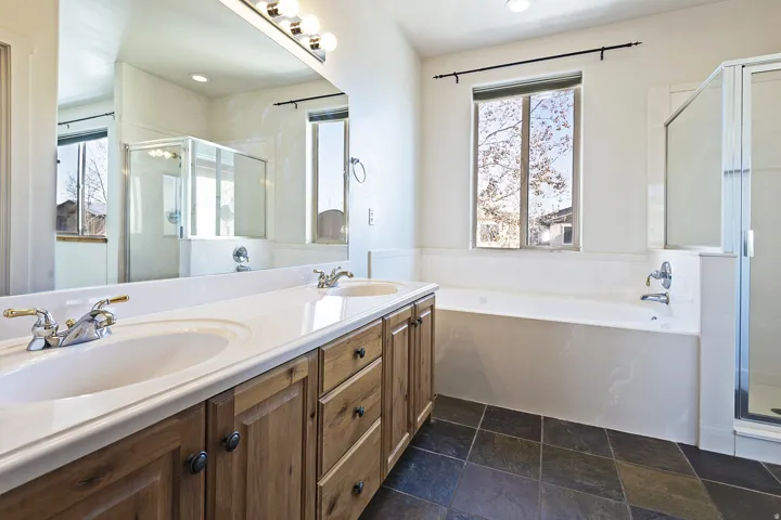 Dual vanity with white countertop, built-in soaking tub, glass-enclosed shower, and dark stone tile flooring