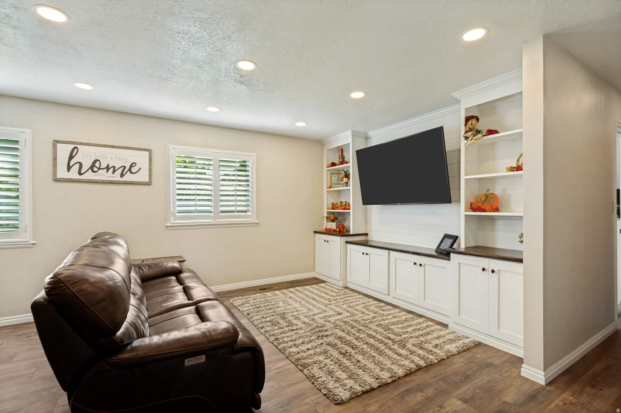 Built-in media console with flanking bookshelves, recessed ceiling lighting, wood-finish flooring, and white plantation shutters