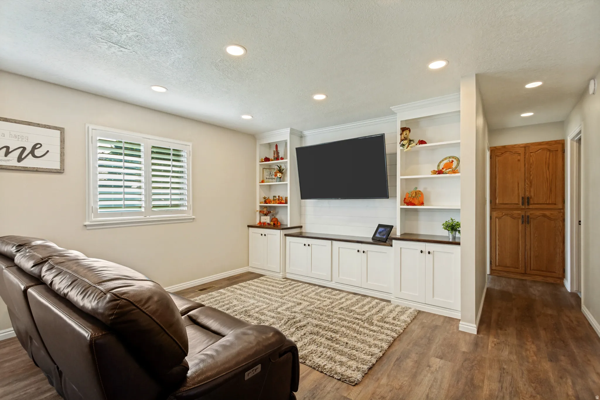 Recessed lighting illuminates the living space featuring wood-finish flooring, a window with plantation shutters, and a built-in media console with open shelving and lower cabinetry