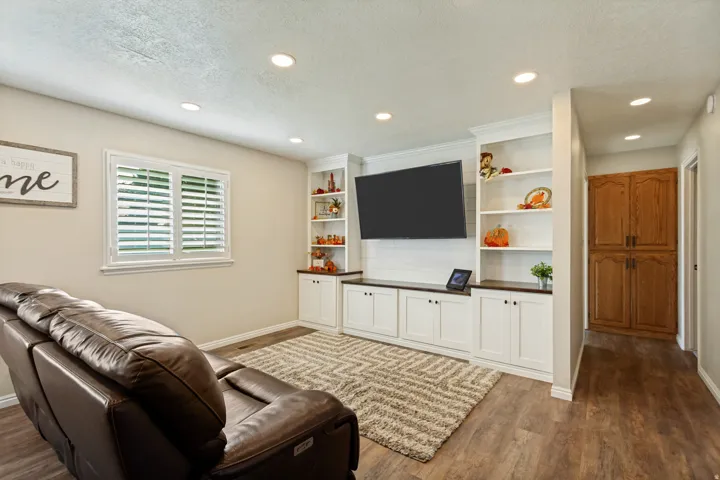 Recessed lighting illuminates the living space featuring wood-finish flooring, a window with plantation shutters, and a built-in media console with open shelving and lower cabinetry