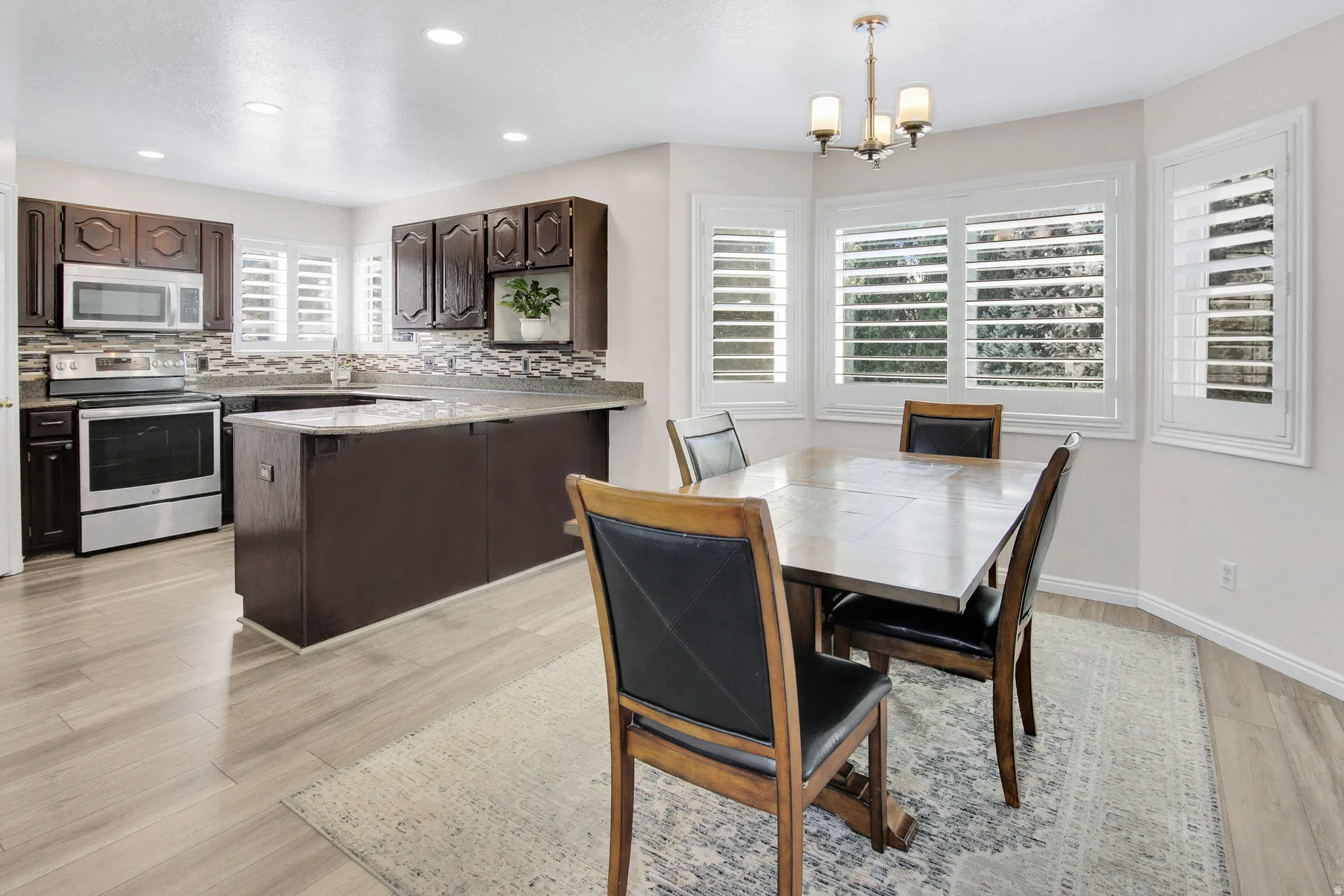 Open-concept kitchen and dining area featuring light wood-finish flooring, dark wood cabinetry, a mosaic tile backsplash, and a built-in stainless steel oven/range with an overhead microwave