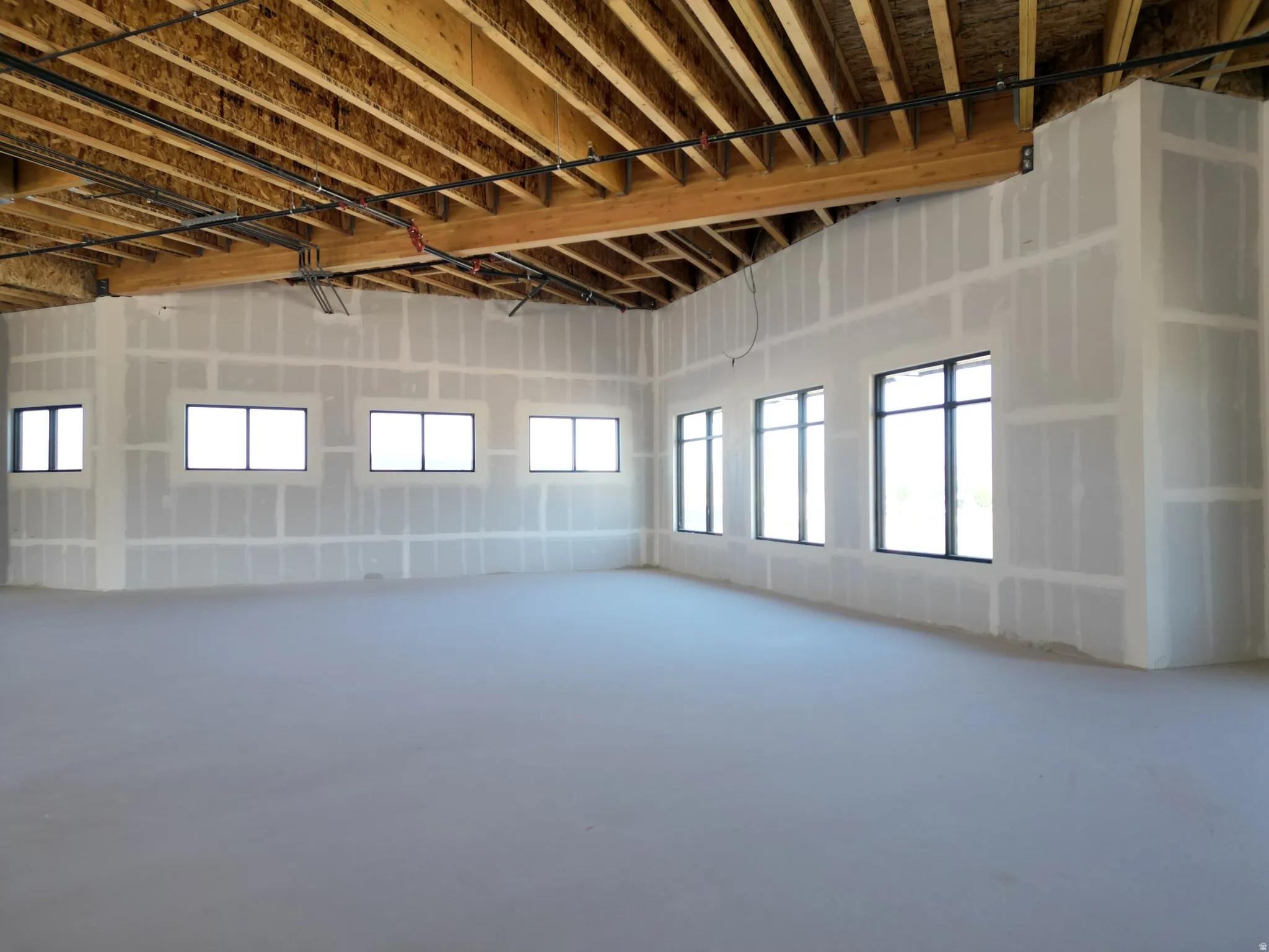 Exposed wood beam ceiling with visible joists, multiple pane windows with dark frames, and unfinished drywall walls