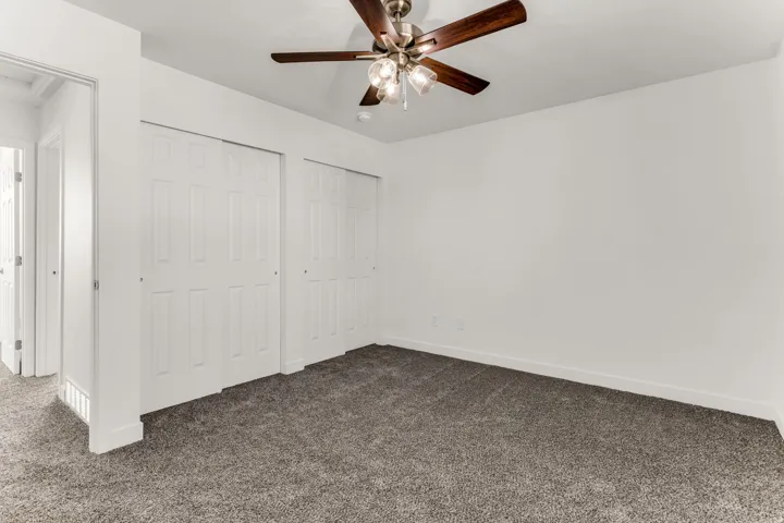 Spacious bedroom with neutral carpet flooring, dual closets with six-panel doors, ceiling fan with integrated lighting, and bright natural light from hallway entry