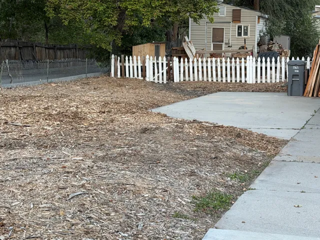 Expansive outdoor space featuring a concrete patio, wood chip ground cover, chain-link fencing, and a white picket fence