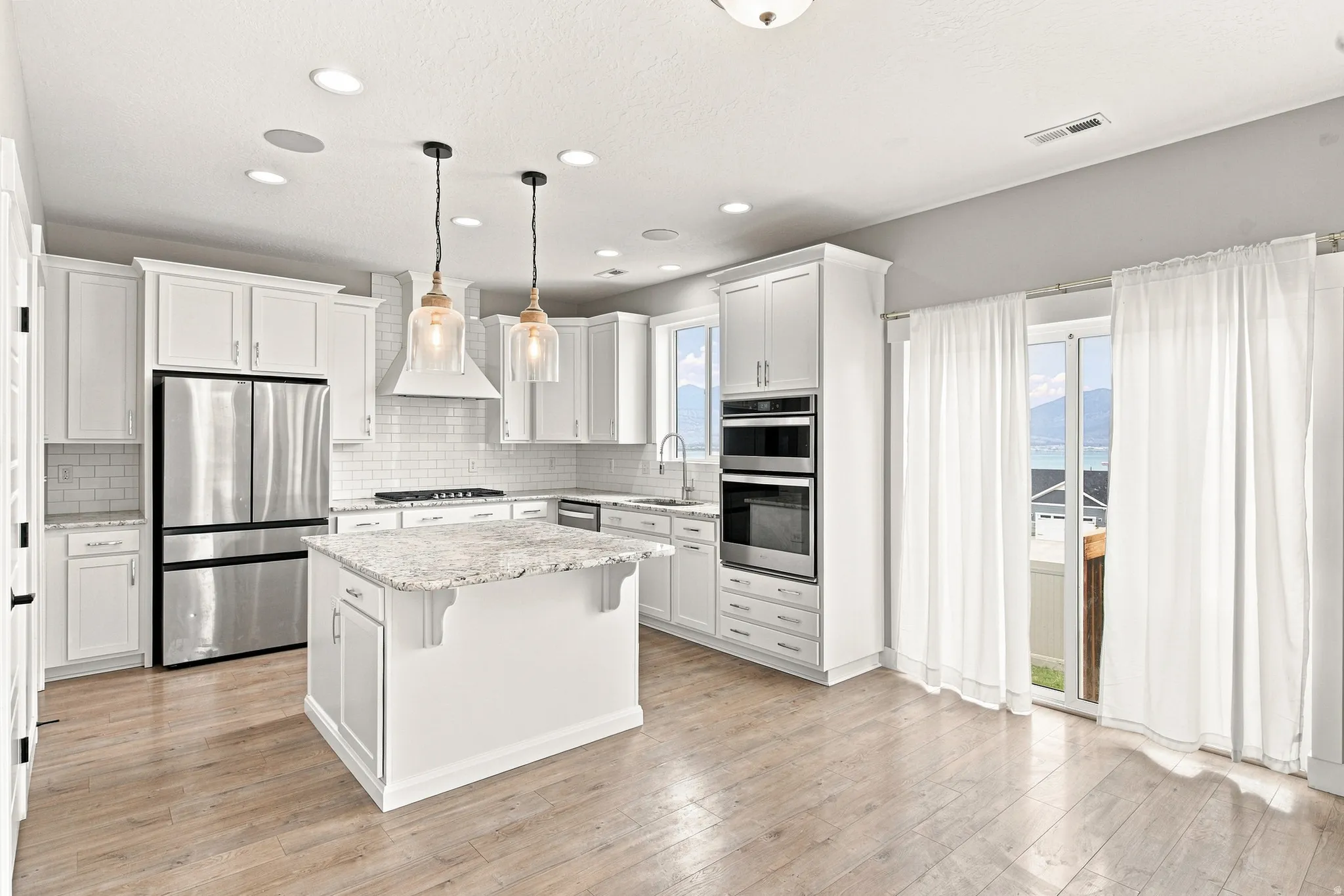 White shaker cabinetry, light-toned wood-finish flooring, stainless steel appliances, central island with stone countertop, and recessed lighting
