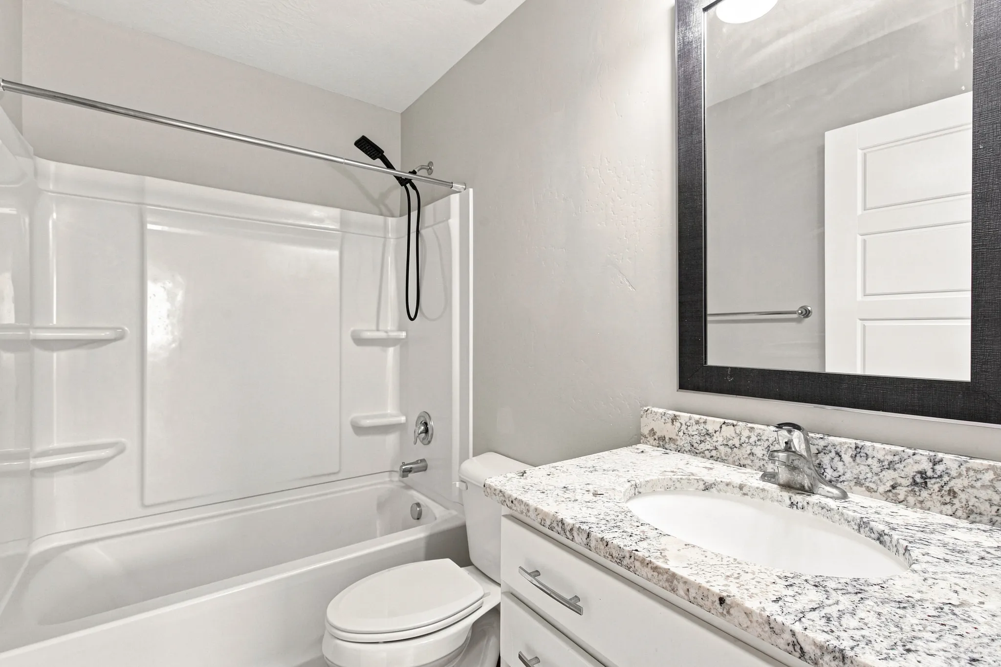 Bathroom featuring a white integrated tub and shower unit, a vanity with a light-colored speckled countertop, an undermount sink, and a framed mirror