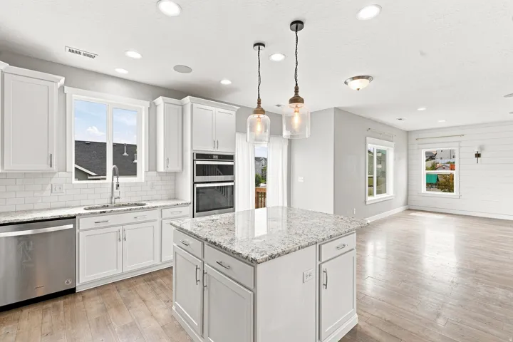 Bright, open-concept kitchen featuring white shaker-style cabinetry, light stone countertops, stainless steel appliances including a double wall oven, and a large center island with seating