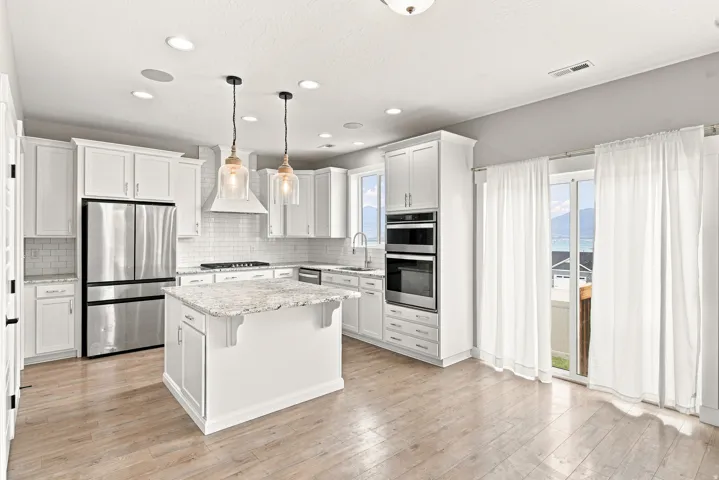 White shaker cabinetry, light-toned wood-finish flooring, stainless steel appliances, central island with stone countertop, and recessed lighting