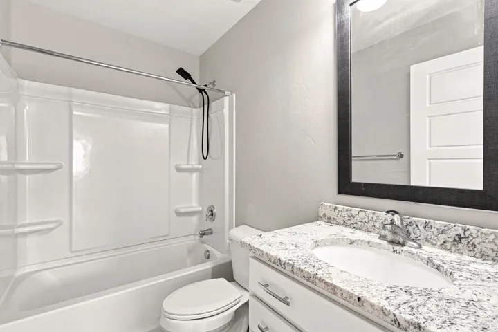 Bathroom featuring a white integrated tub and shower unit, a vanity with a light-colored speckled countertop, an undermount sink, and a framed mirror