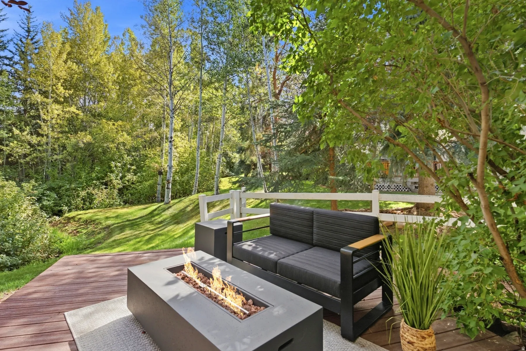 Expansive wood-finish decking with an integrated fire feature, set against a backdrop of mature trees and a sloped lawn