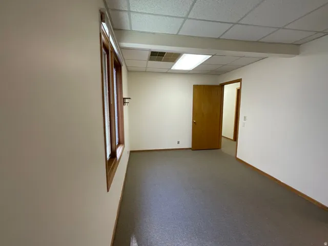 Interior room with wood-trimmed window, neutral wall paint, suspended acoustical ceiling, and grey carpeting