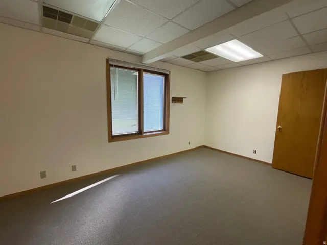 Neutral-toned wall paint, drop ceiling with integrated lighting, wood-trimmed window with blinds, carpeted flooring, and a wood-paneled door