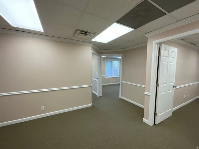 Neutral-toned walls with chair rail molding, drop ceiling with integrated fluorescent lighting, and carpeted flooring throughout