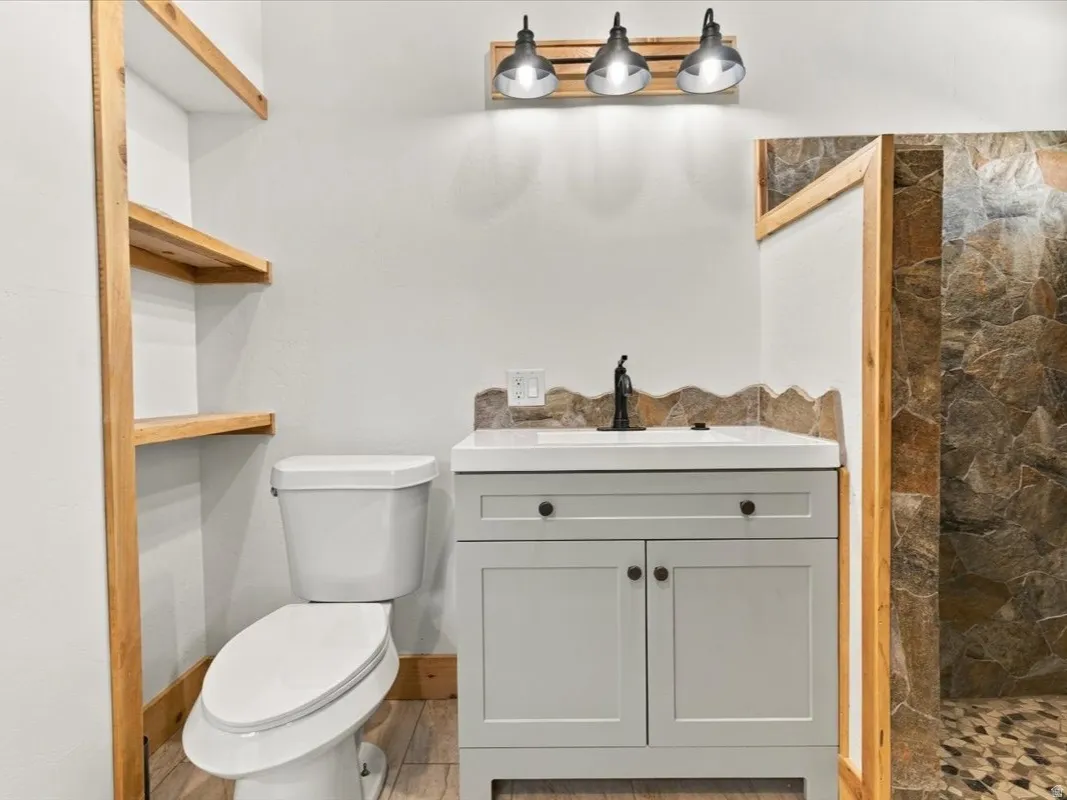 Bathroom featuring a white vanity with integrated sink, open wood shelving, a toilet, a shower with stone tile walls, and a pebble-finish shower floor