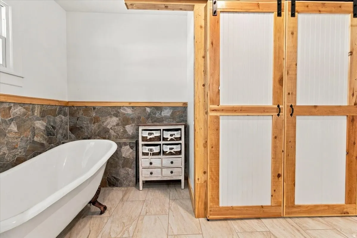 Freestanding soaking tub, stone tile wainscoting, wood-finish tile flooring, and barn-style sliding doors