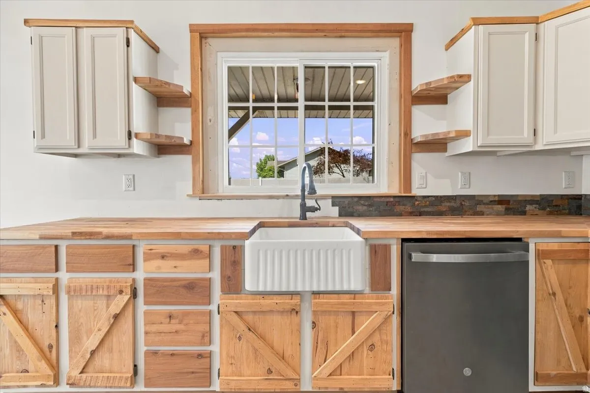 Wood-finish countertops, white farmhouse sink with a gooseneck faucet, stainless steel dishwasher, wood-finish lower cabinetry with barn door accents, and upper white shaker cabinetry