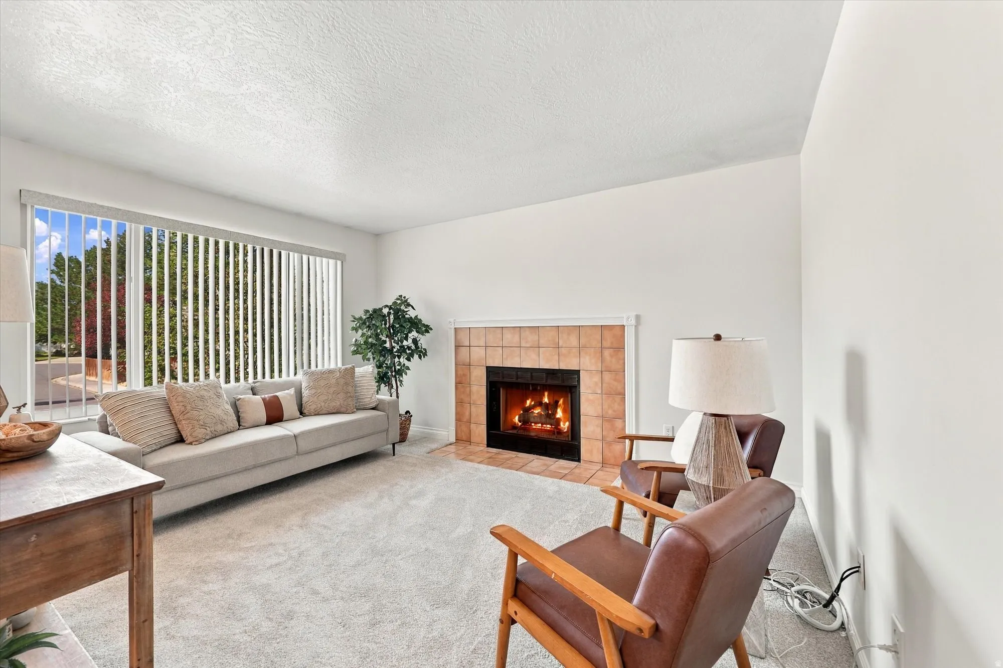 Living area with a tiled fireplace, wide window with vertical blinds, and light-colored carpeting