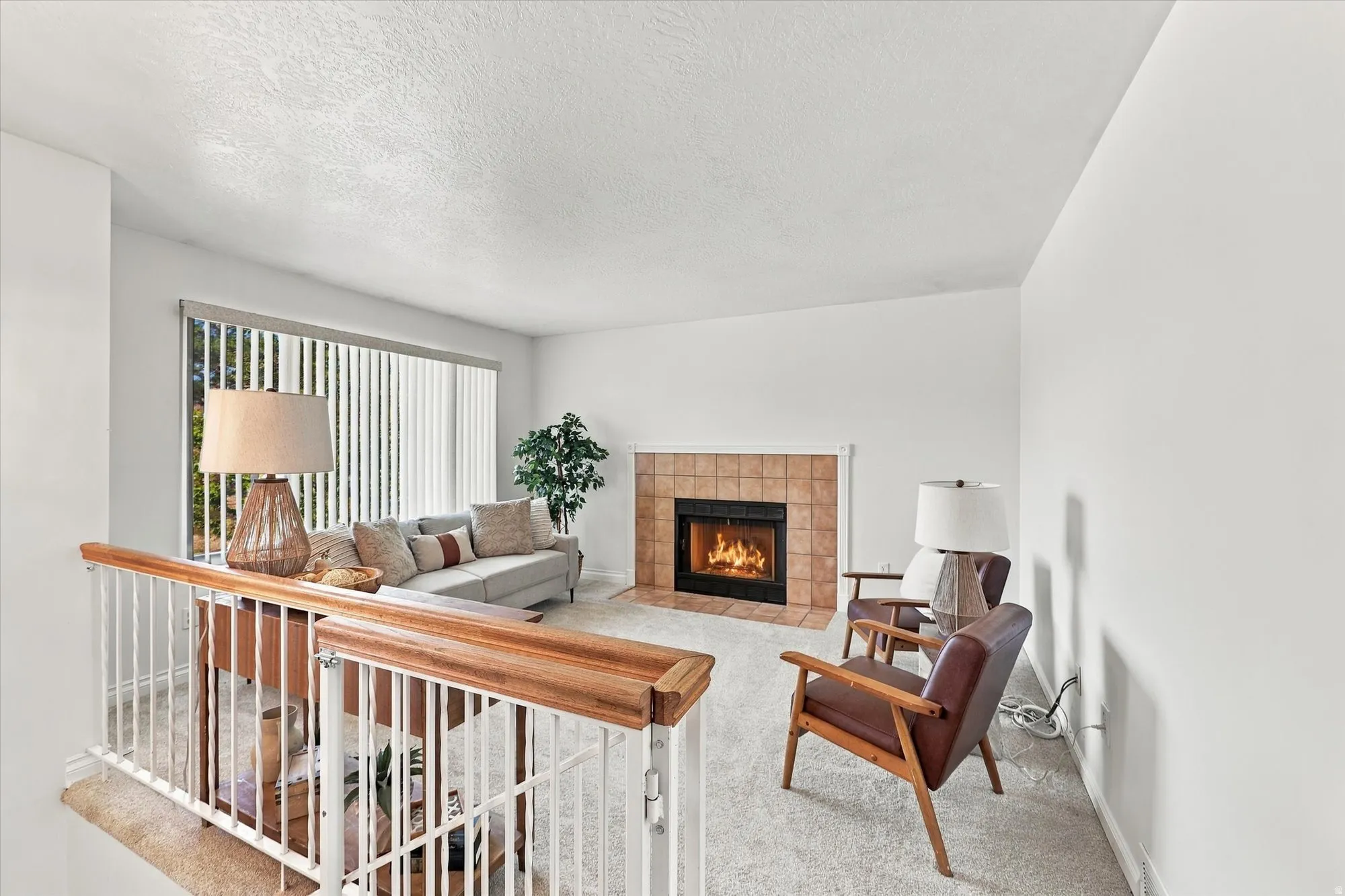 Living space featuring a tile-surround fireplace, large windows with vertical blinds, and light-toned carpeting