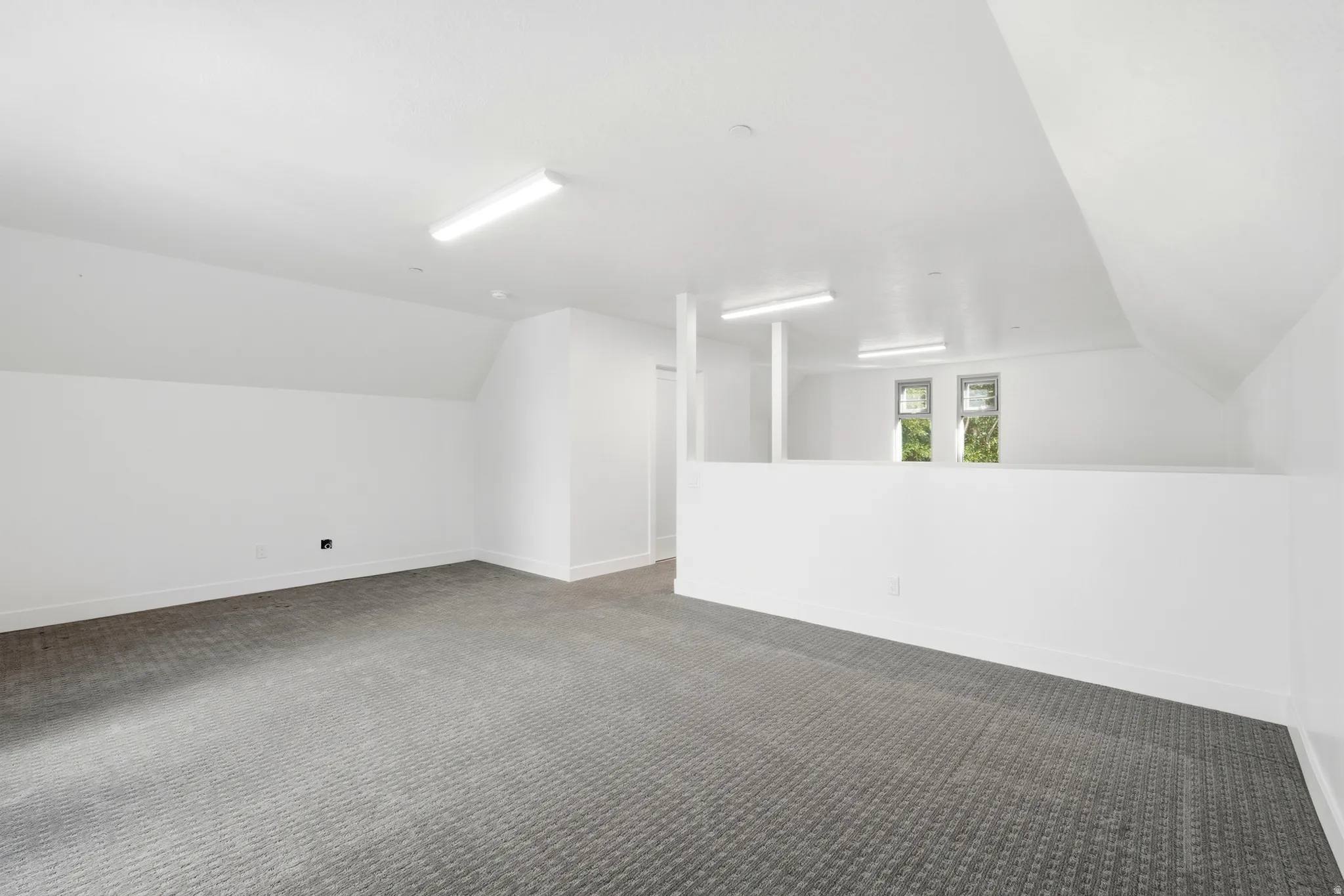 Expansive room featuring clean white walls, grey patterned carpeting, a partial wall, and multiple windows providing natural light