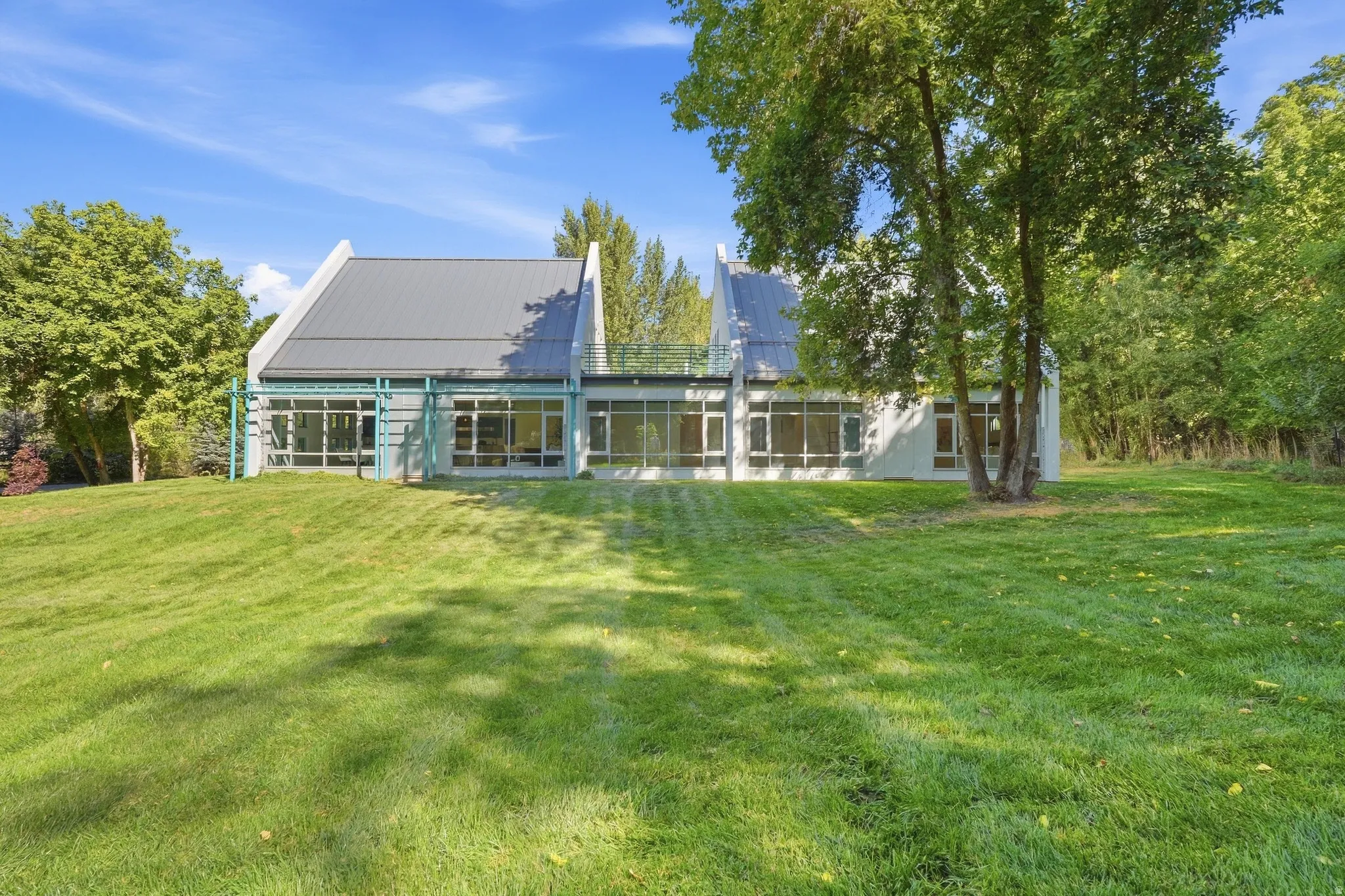 Modern architectural design featuring a sloped metal roof, extensive floor-to-ceiling windows, and a surrounding expansive lawn with mature trees