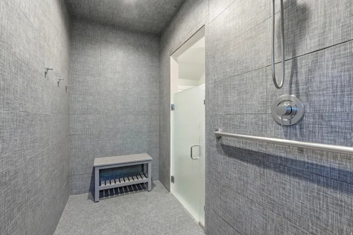 Textured gray wall tiling throughout, brushed nickel shower fixtures, and a frosted glass shower door