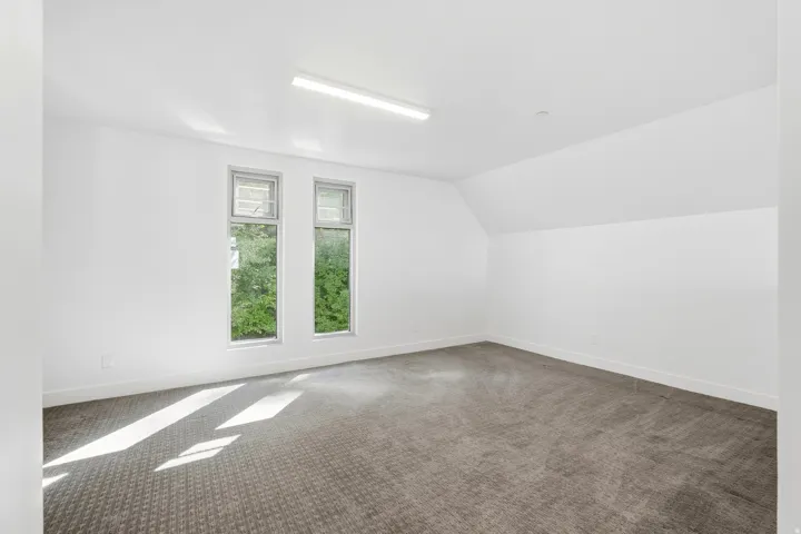 Spacious room with sloped ceilings, two vertical windows, textured carpet, and a fluorescent light fixture
