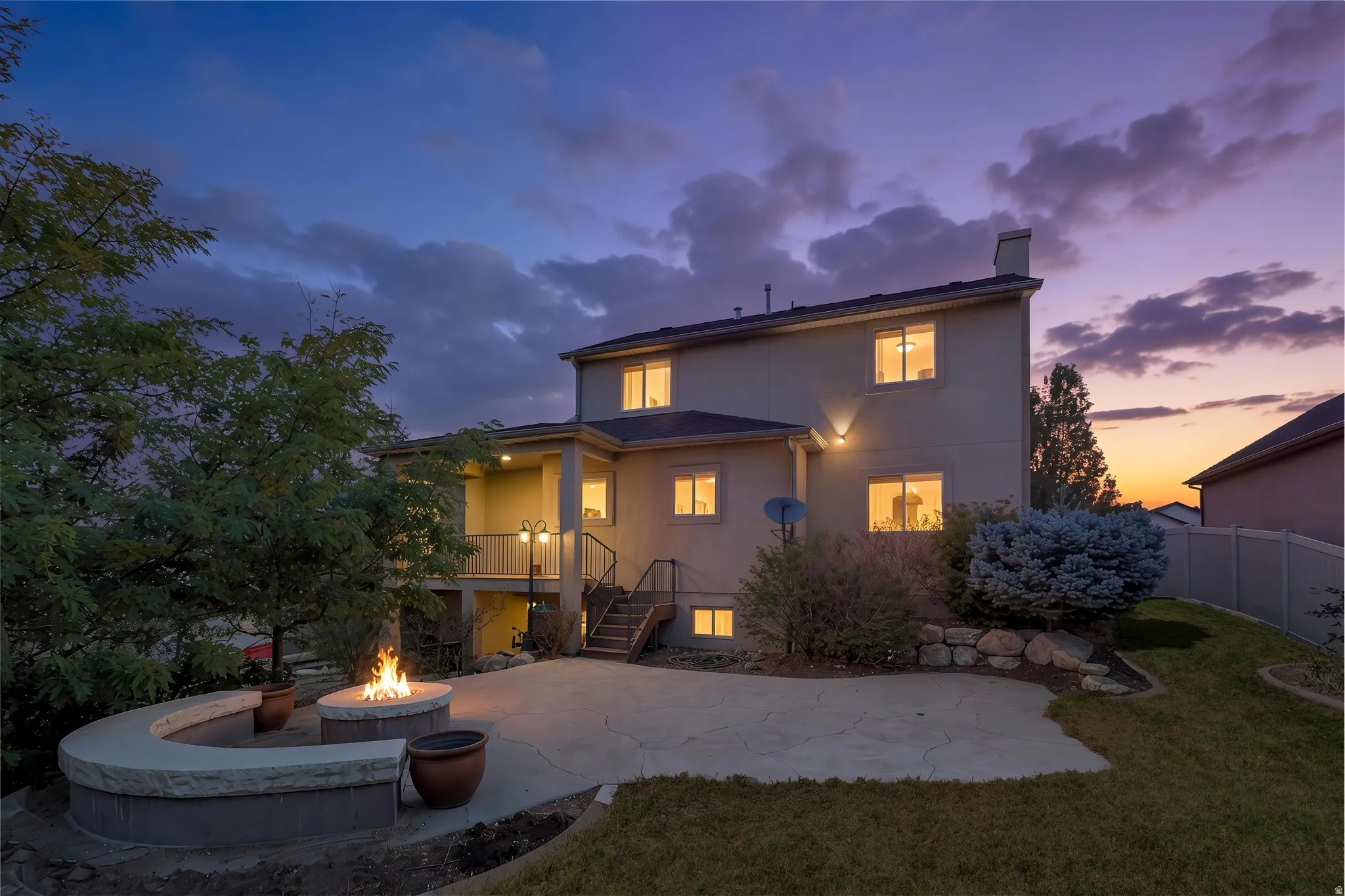 Rear exterior showcasing a built-in fire pit with stone seating, concrete patio, and a covered balcony, twilight added