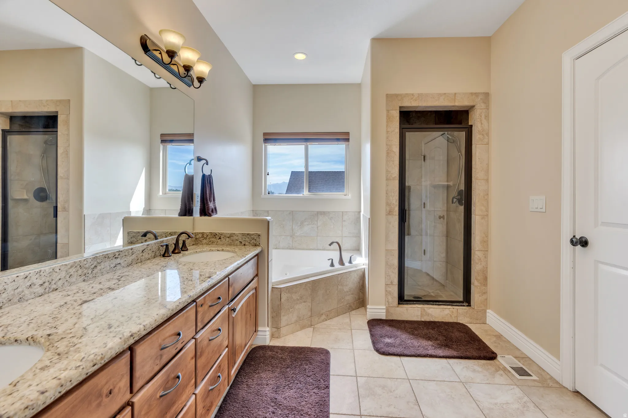 Dual vanity with light-toned countertop, wood-finish cabinetry, tile flooring, corner soaking tub, and a separate glass-enclosed shower