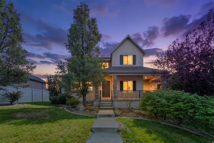 Gabled roofline with decorative shutters, stone-accented columns, covered front porch with railing, mature trees, and a paved walkway, twilight added