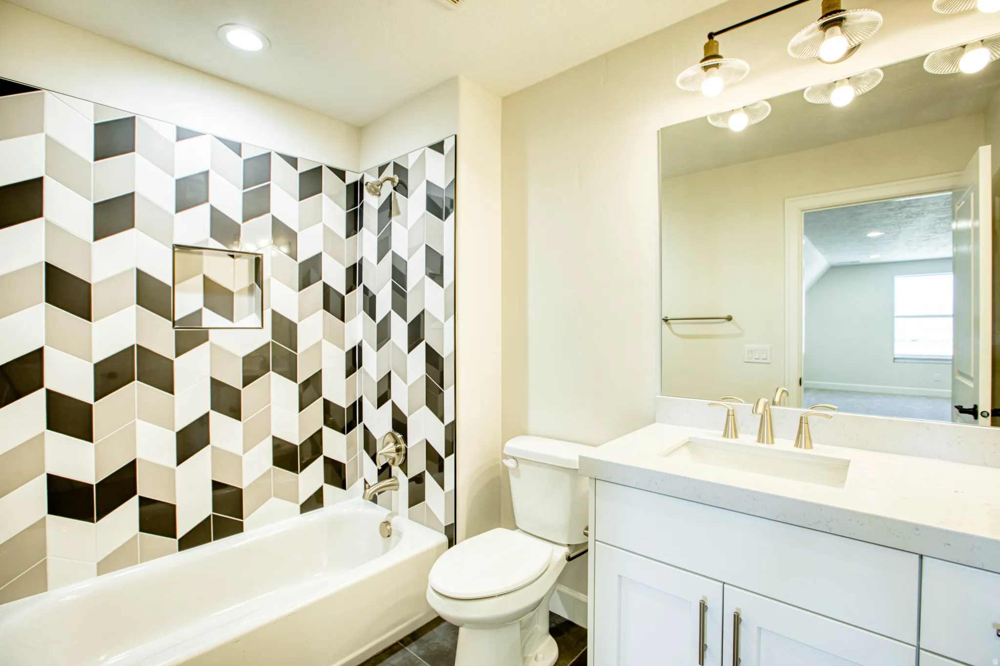 Bathtub with chevron-patterned tile surround and recessed niche, single vanity with light-toned countertop, undermount sink, and brushed nickel faucet, wall-mounted light fixture with clear glass shades