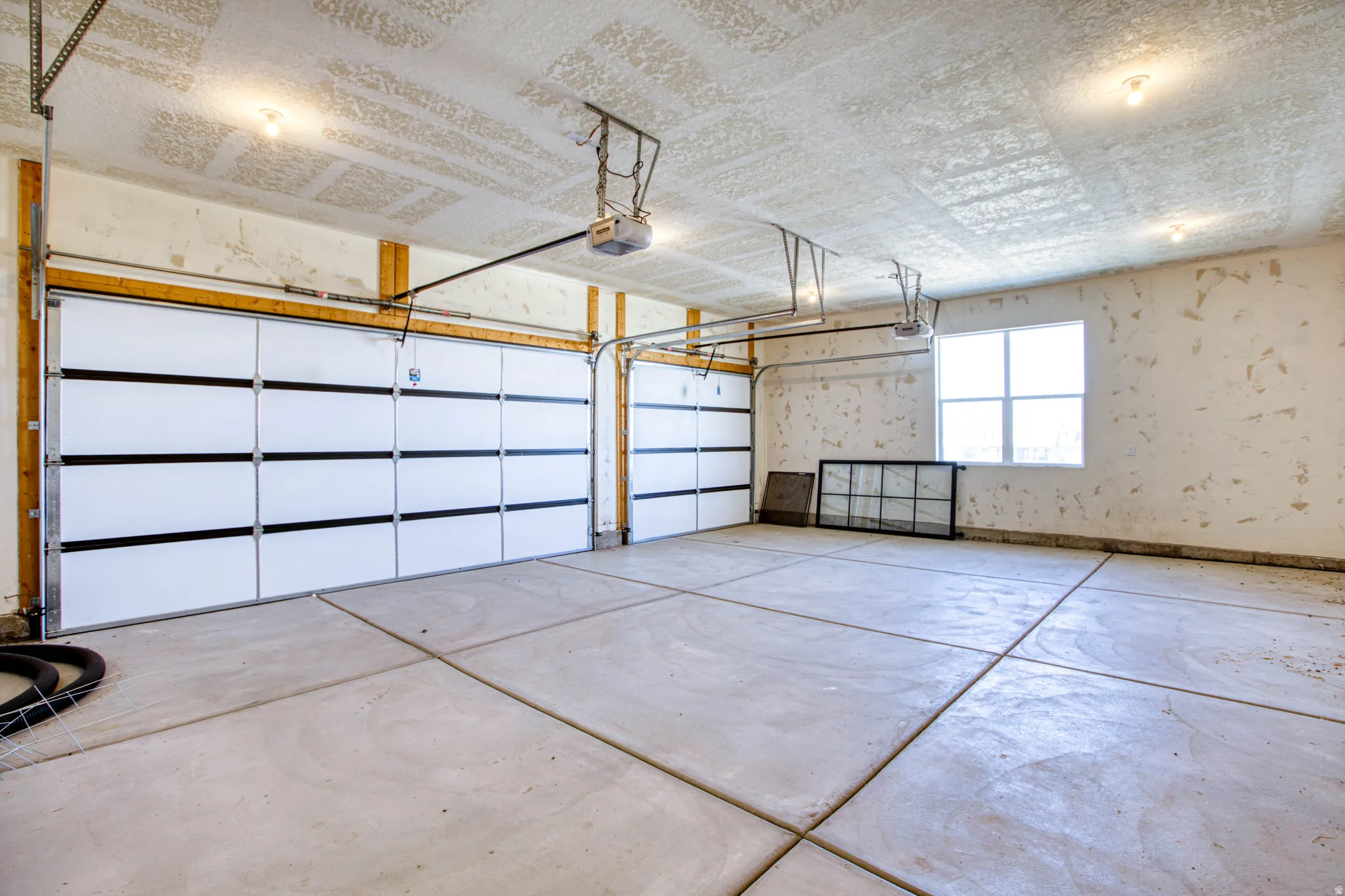Two white garage doors with black horizontal accents, concrete flooring with expansion joints, a four-pane window, and exposed ceiling joists