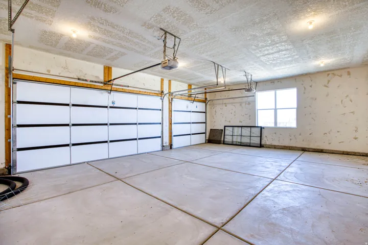 Two white garage doors with black horizontal accents, concrete flooring with expansion joints, a four-pane window, and exposed ceiling joists