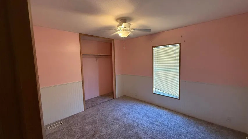 Room featuring carpeting, a built-in closet with rod, a window with blinds, a ceiling fan with light fixture, and wainscoting
