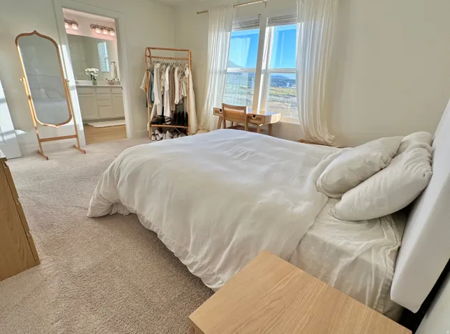 Plush carpet flooring, large windows with white curtains, and a light-colored headboard