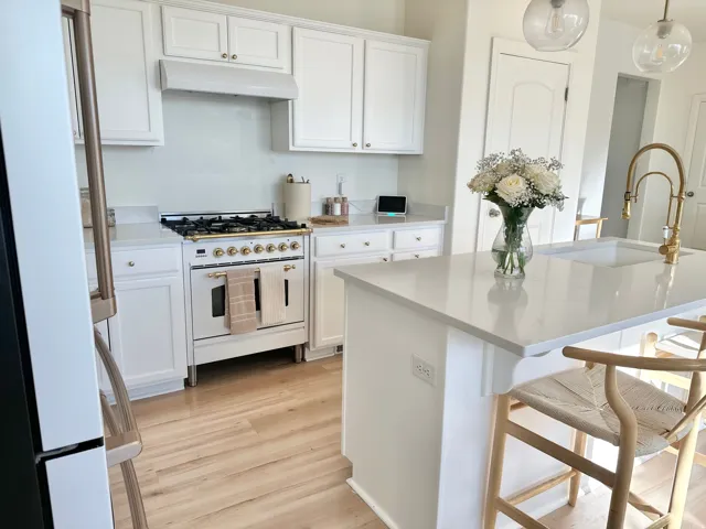 White cabinetry, light wood-finish flooring, white countertops, kitchen island with integrated sink, and brass-tone fixtures
