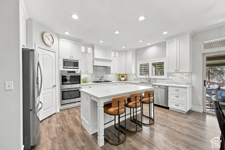 Kitchen featuring white cabinetry, wood-finish flooring, stainless steel appliances, a center island, and a tile backsplash