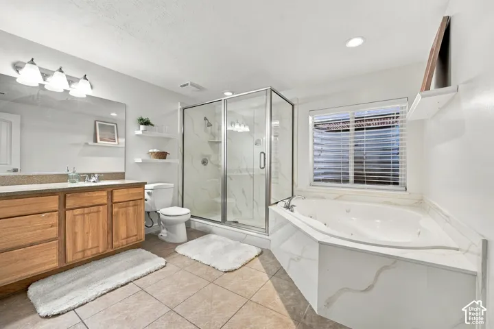 Double vanity with wood-finish cabinetry, corner soaking tub, walk-in shower with glass enclosure, white floating shelves, and tile flooring